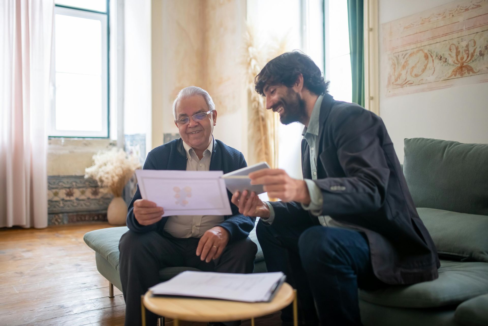 An older man and a younger man looking at paperwork on a couch in a living room. Both are smiling, with natural light from a window.