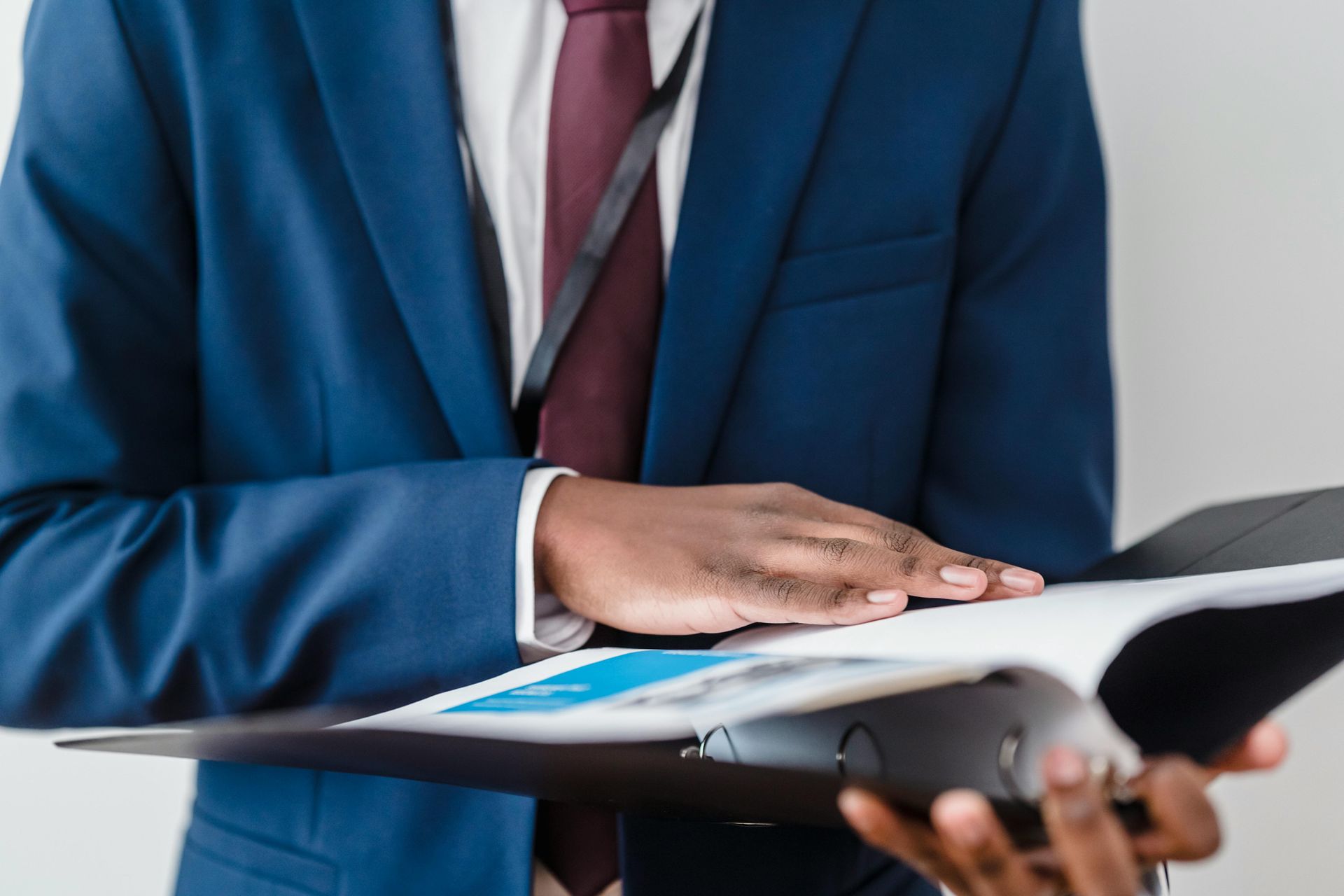 Person in a blue suit examines documents in a black binder. They are wearing a burgundy tie and a lanyard.
