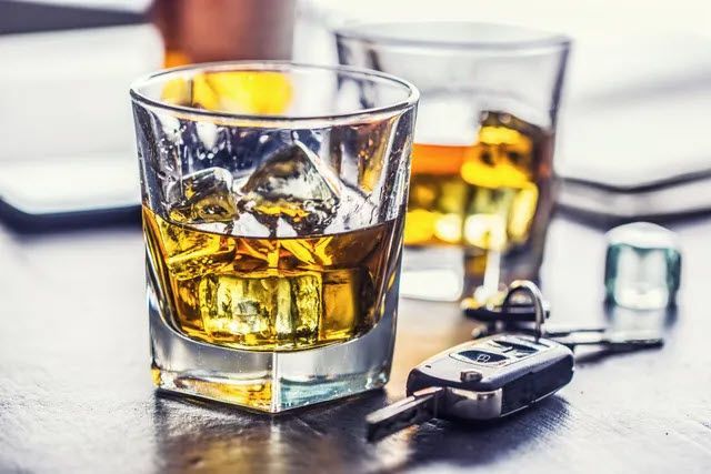 Whiskey in a rocks glass with ice cubes, car keys on a table near other drinks, representing drunk driving.