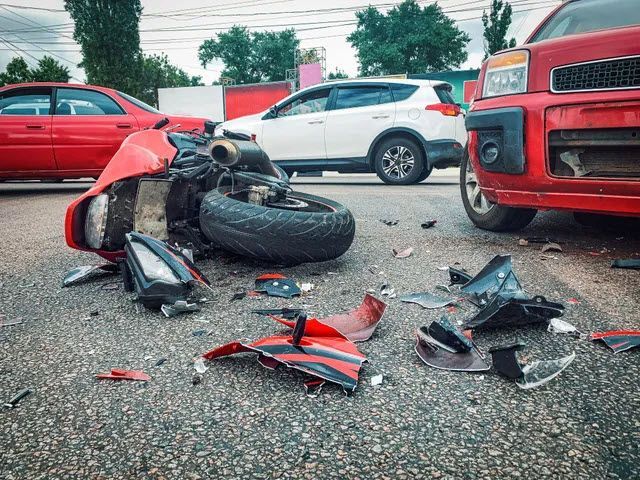 Motorcycle wreckage on asphalt after a collision with red and white cars. Debris scattered.