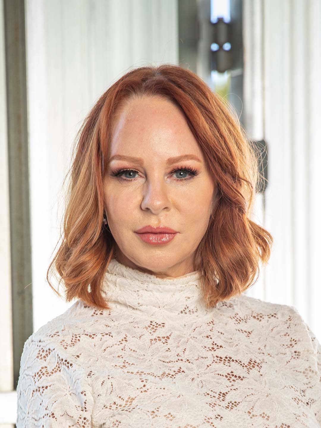 A woman with red hair and blue eyes is wearing a white lace sweater.