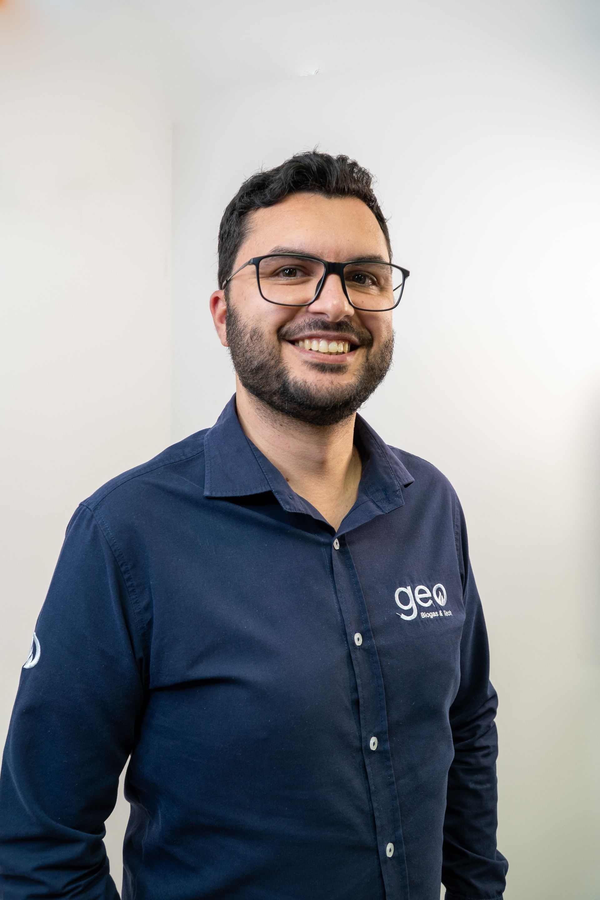Um homem com barba e óculos está vestindo uma camisa azul e sorrindo.