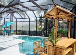 A swimming pool inside a screened-in lanai with a bamboo tiki bar and a small table with chairs under a blue umbrella.