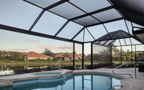 A swimming pool and hot tub inside a screened lanai, overlooking a lake and suburban homes at sunset.
