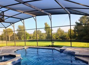 A blue swimming pool with a hot tub and water feature enclosed in a screened lanai overlooking a grassy lawn and trees.