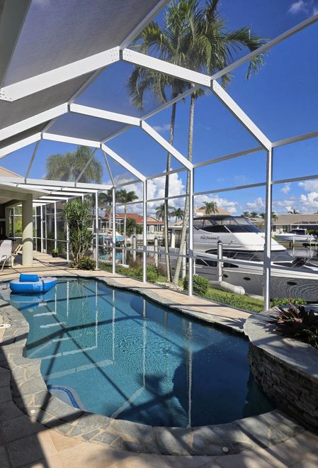A swimming pool inside a screened-in lanai overlooking a canal with a parked yacht and palm trees on a sunny day.