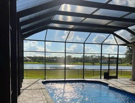 A backyard swimming pool enclosed in a dark metal screen cage, overlooking a lake and grassy landscape under a blue sky.