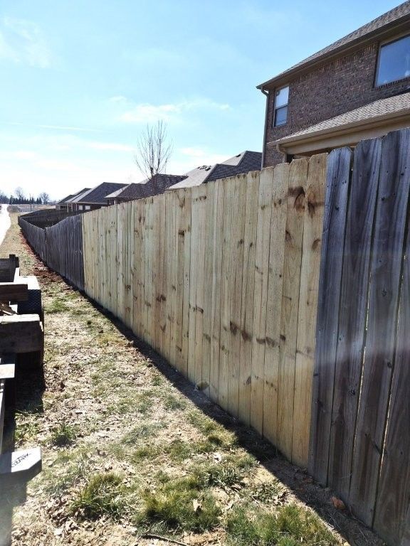 A new, light-colored wooden fence section stands prominently in front of older, weathered gray fencing along a backyard.