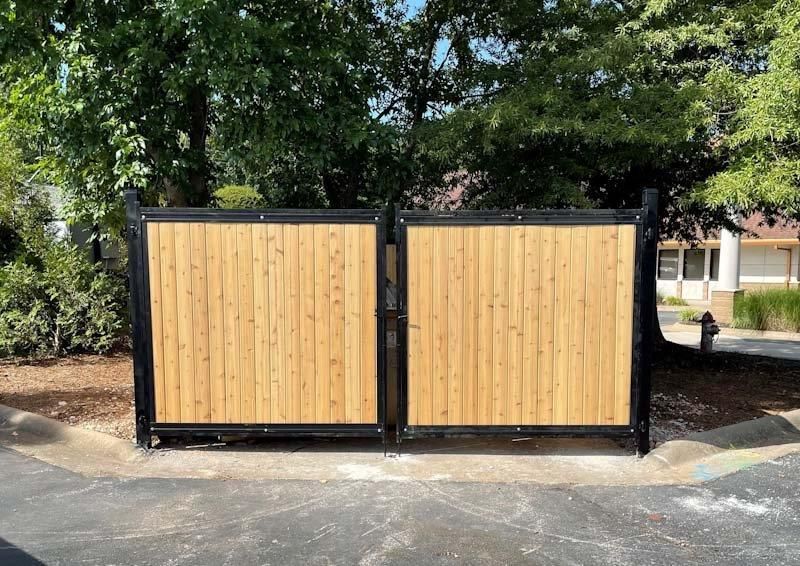 A double wooden gate with vertical slats and a black metal frame stands outdoors on a paved surface near trees.