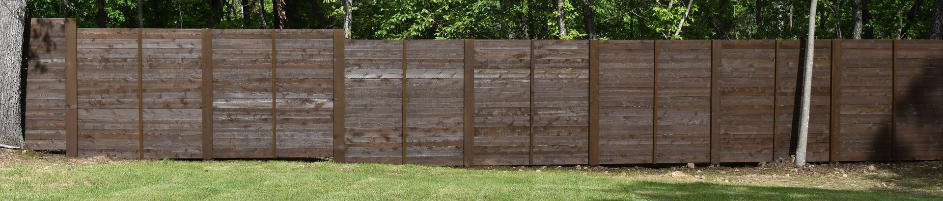 A long, horizontal wooden privacy fence made of dark, horizontal planks with vertical posts against a backdrop of trees.