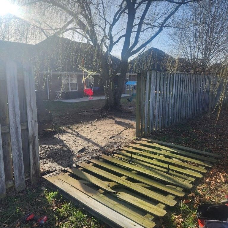 A wooden fence gate lies broken and detached on the ground in a backyard, revealing an opening between two fence sections.