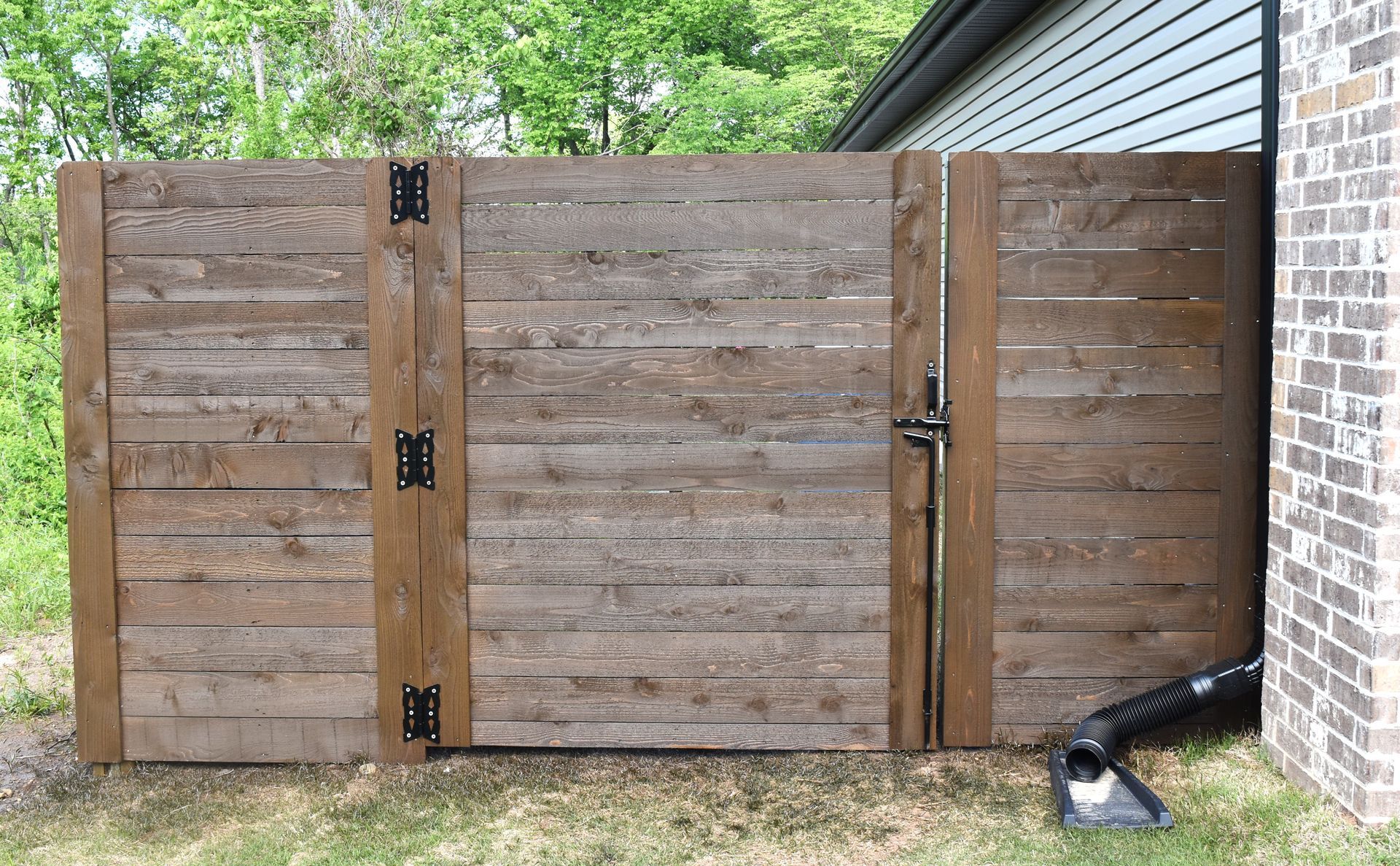 A wooden privacy fence gate attached to a brick house wall, featuring horizontal slats and black metal hinges and latches.