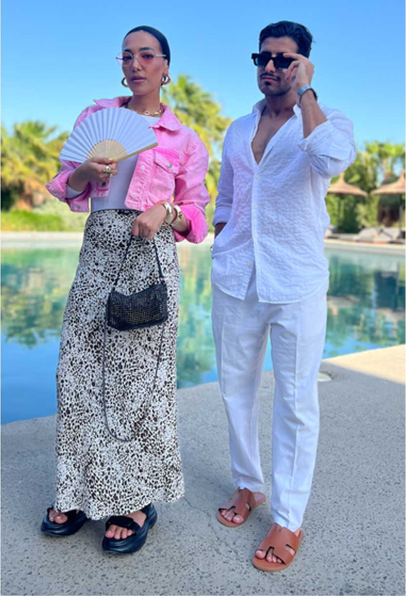 Woman in pink jacket and patterned skirt holds a fan next to a man in a white shirt and pants; by a pool.