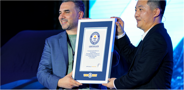 Two men holding a framed certificate, one man smiles. Dark background with blue lighting.