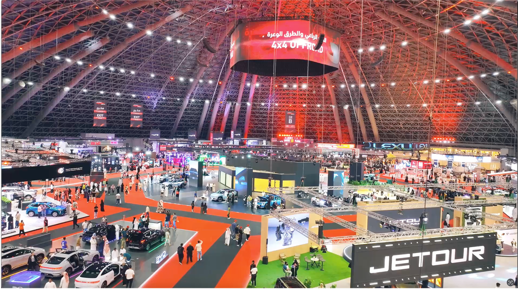 Large, crowded auto show in a domed hall, with cars on display and people viewing exhibits.