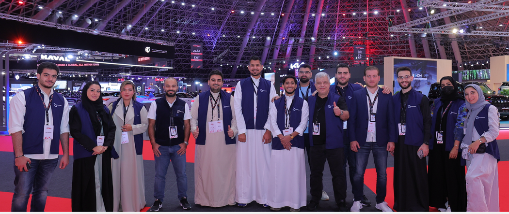 A group of people wearing vests in a large venue. Some are wearing traditional white robes. Purple and red lighting.