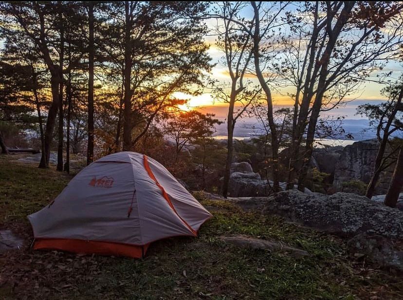 Camping tent on a hilltop at sunset. Orange and gray tent, trees, and rocky landscape.