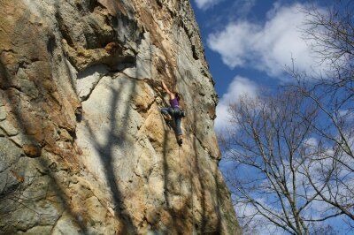 Rock climber ascending a textured, sunlit cliff face. Blue sky, bare trees.