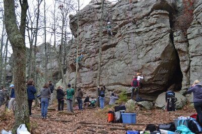 Rock climbers on a large rock face; several people watch and set up equipment in a wooded area.