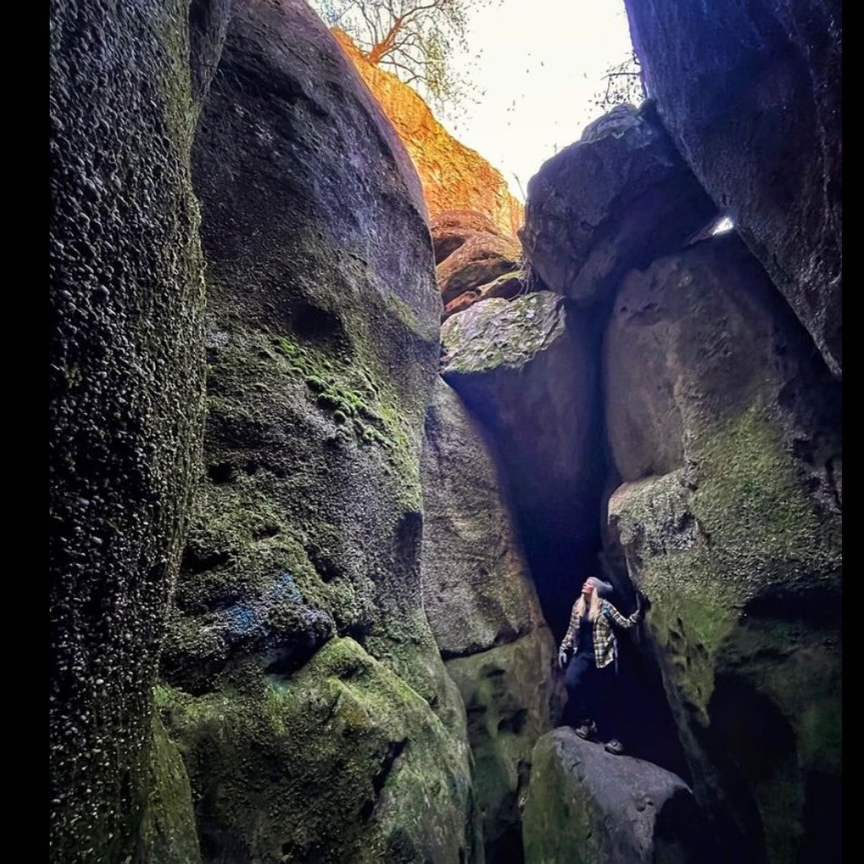 Person stands inside a narrow rock crevice; moss covers stone walls. Sunlight illuminates the opening.