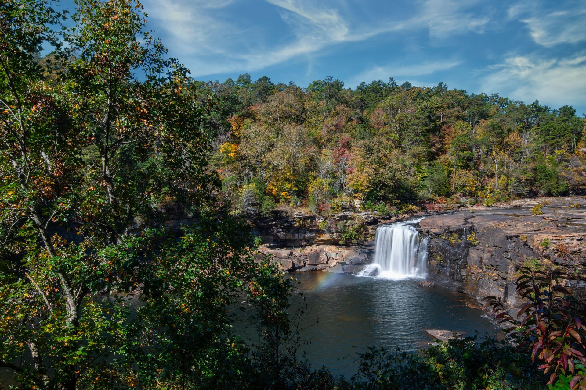 Waterfall cascading into a calm pool, surrounded by autumn foliage and a blue sky.