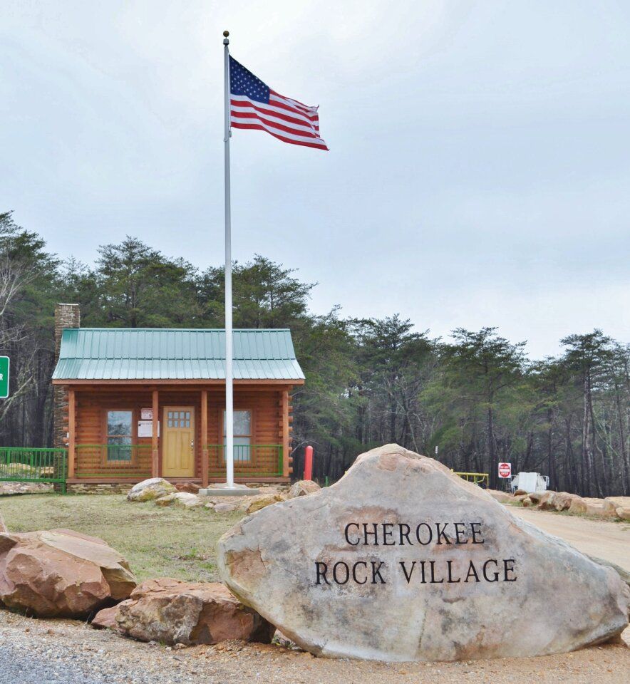 Cherokee Rock Village sign, log cabin, and American flag on a pole under cloudy sky.
