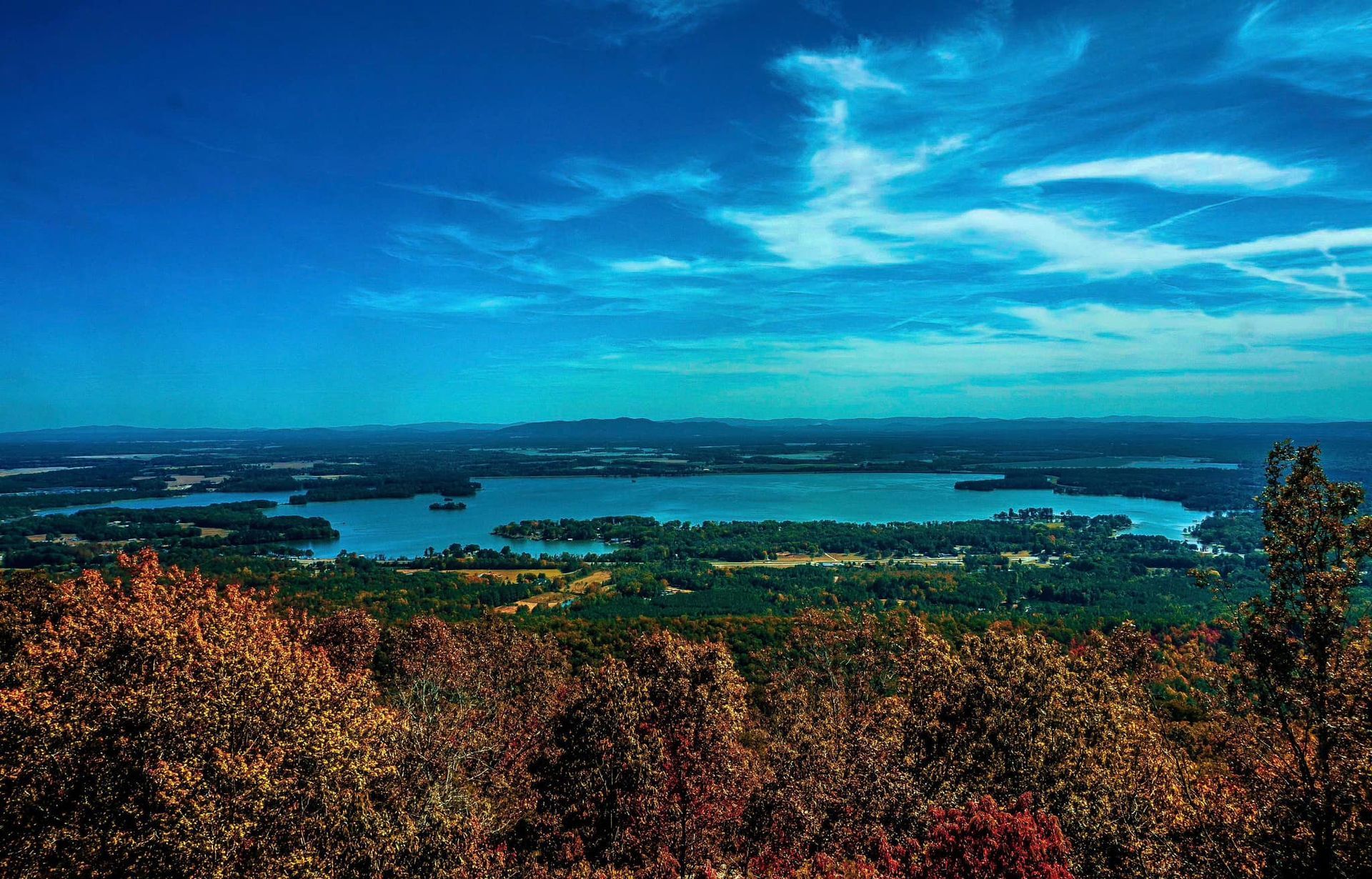 View from a mountaintop overlooking a lake, trees, and blue sky with clouds.