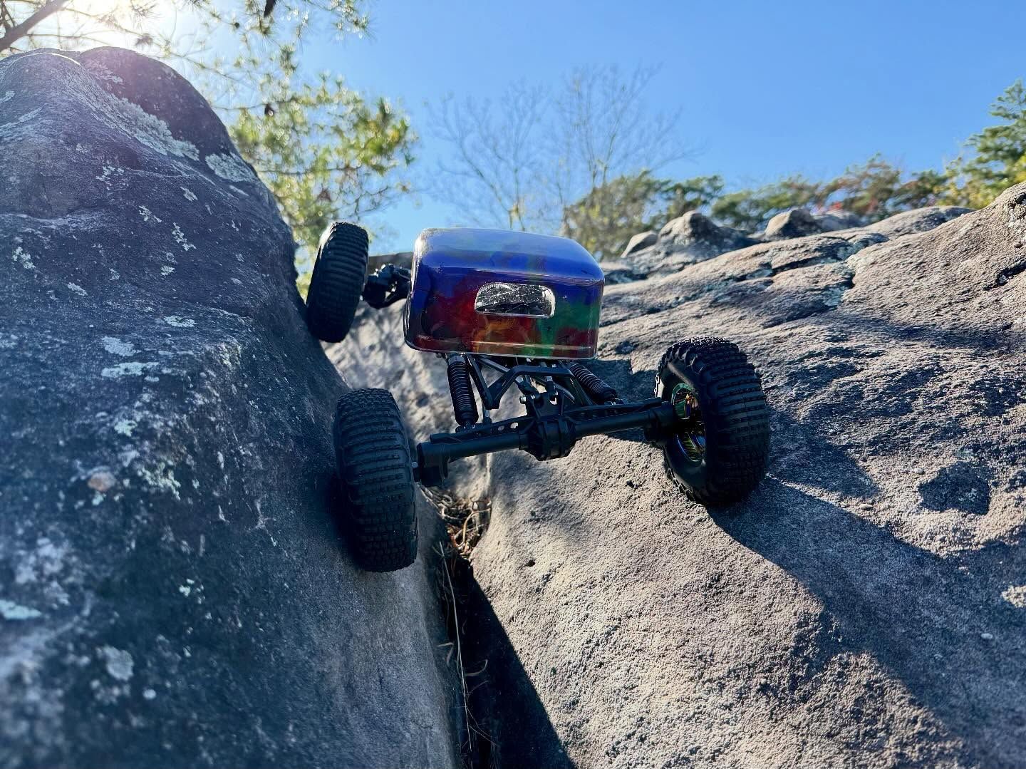 RC car climbs a rocky cliff with a blue and rainbow colored body under a blue sky.