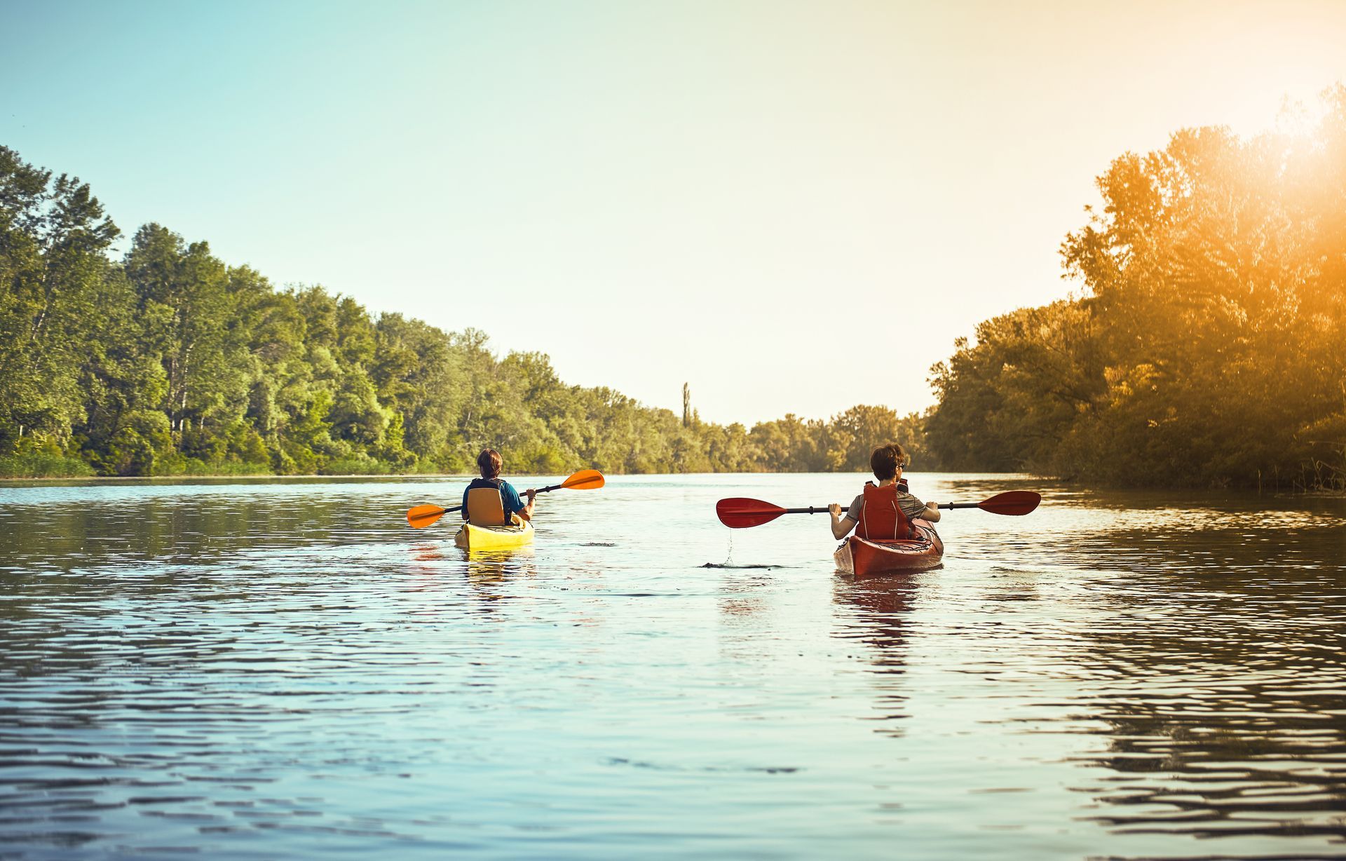 Two people kayaking on a calm river, sunny day, trees on either side.