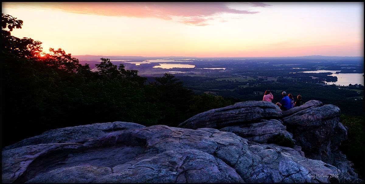 Sunset view from rocky mountaintop, overlooking landscape and water. Several people sit, silhouetted.