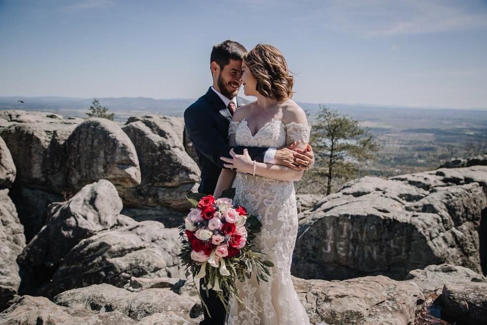 Bride and groom embrace on a rocky mountaintop, holding a bouquet, with a scenic valley view.