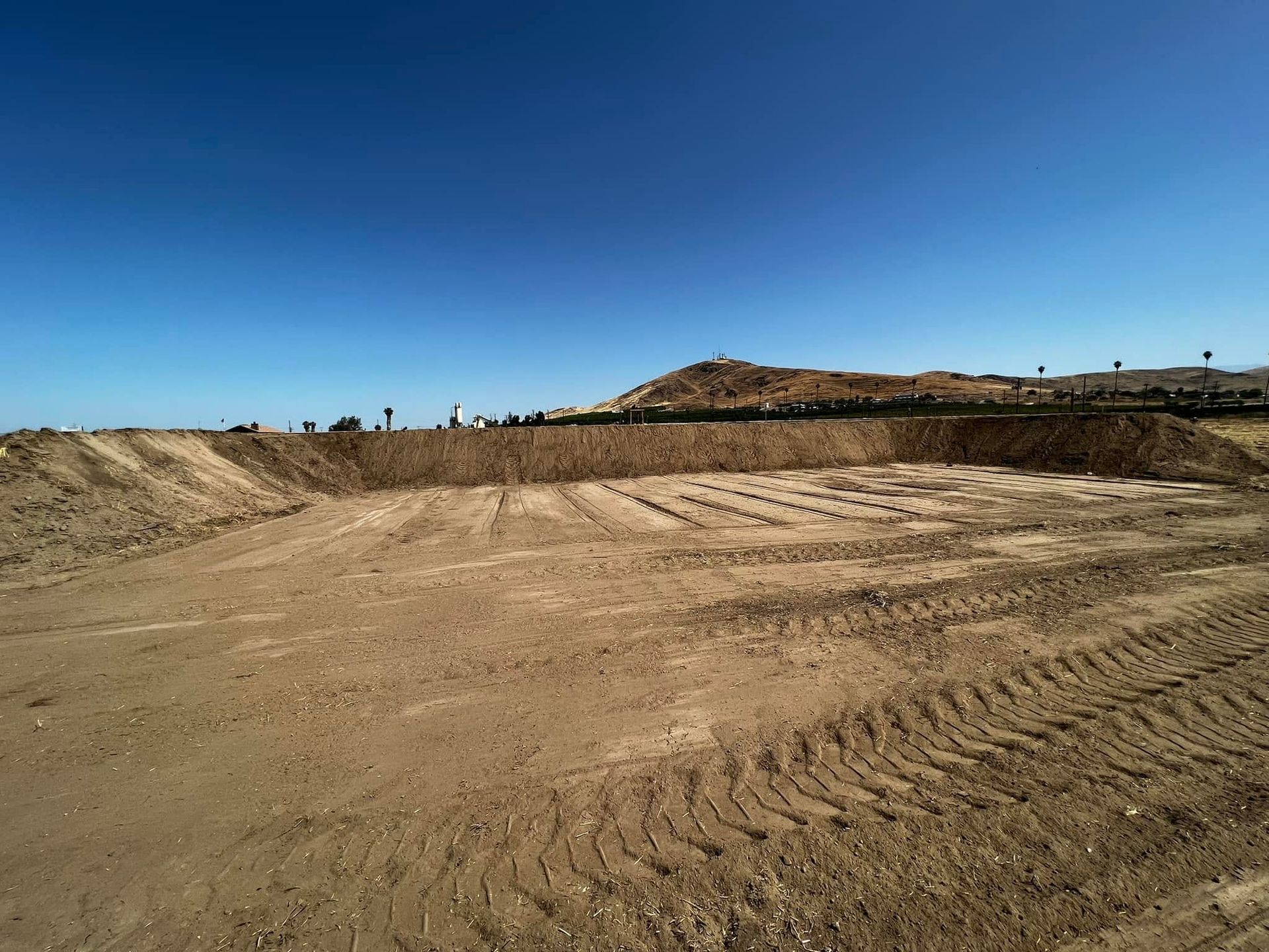 Cleared dirt lot with tire tracks under a clear blue sky, low hills in the distance