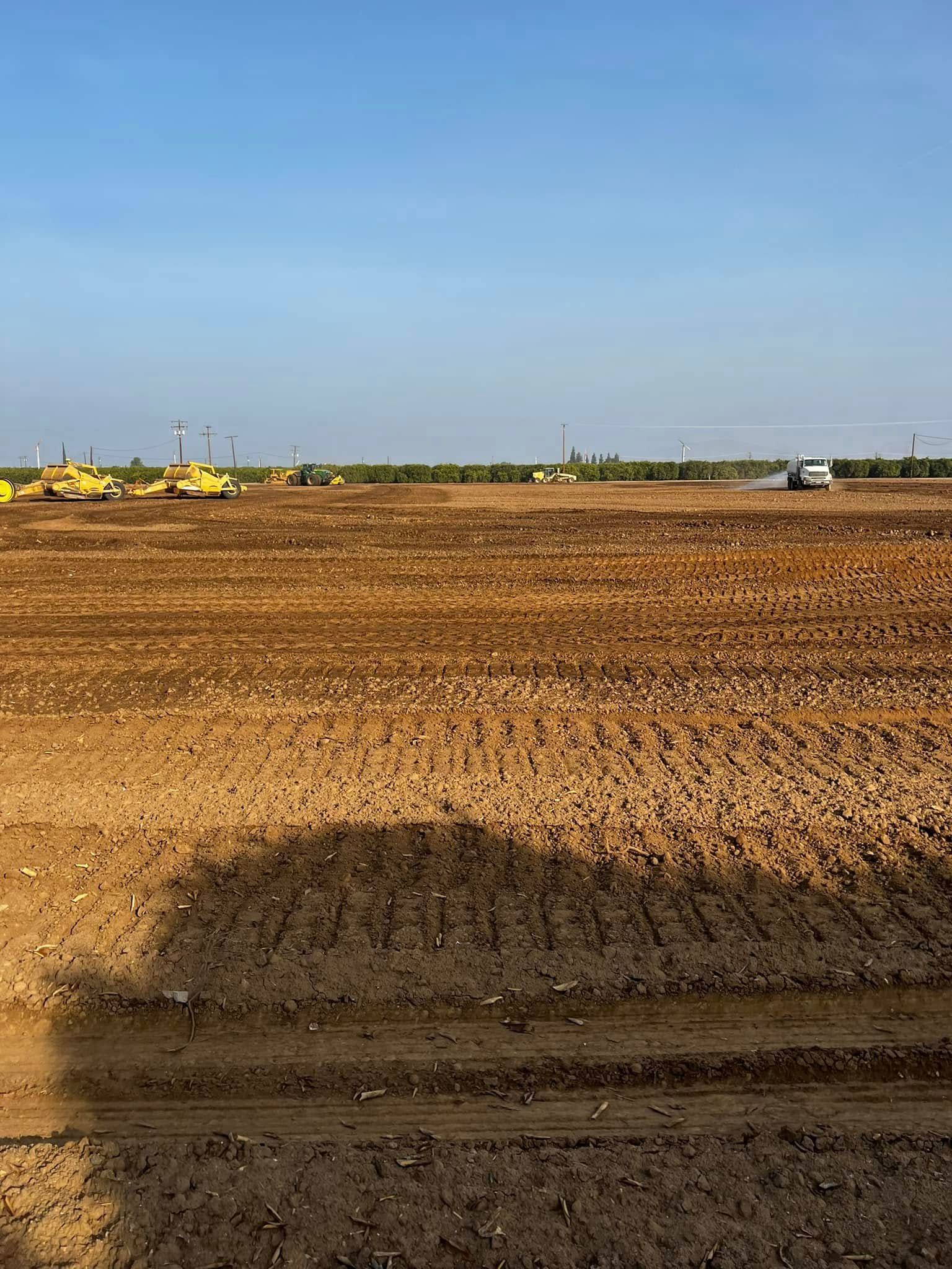 Plowed brown field under a clear blue sky, with machinery on the distant horizon.