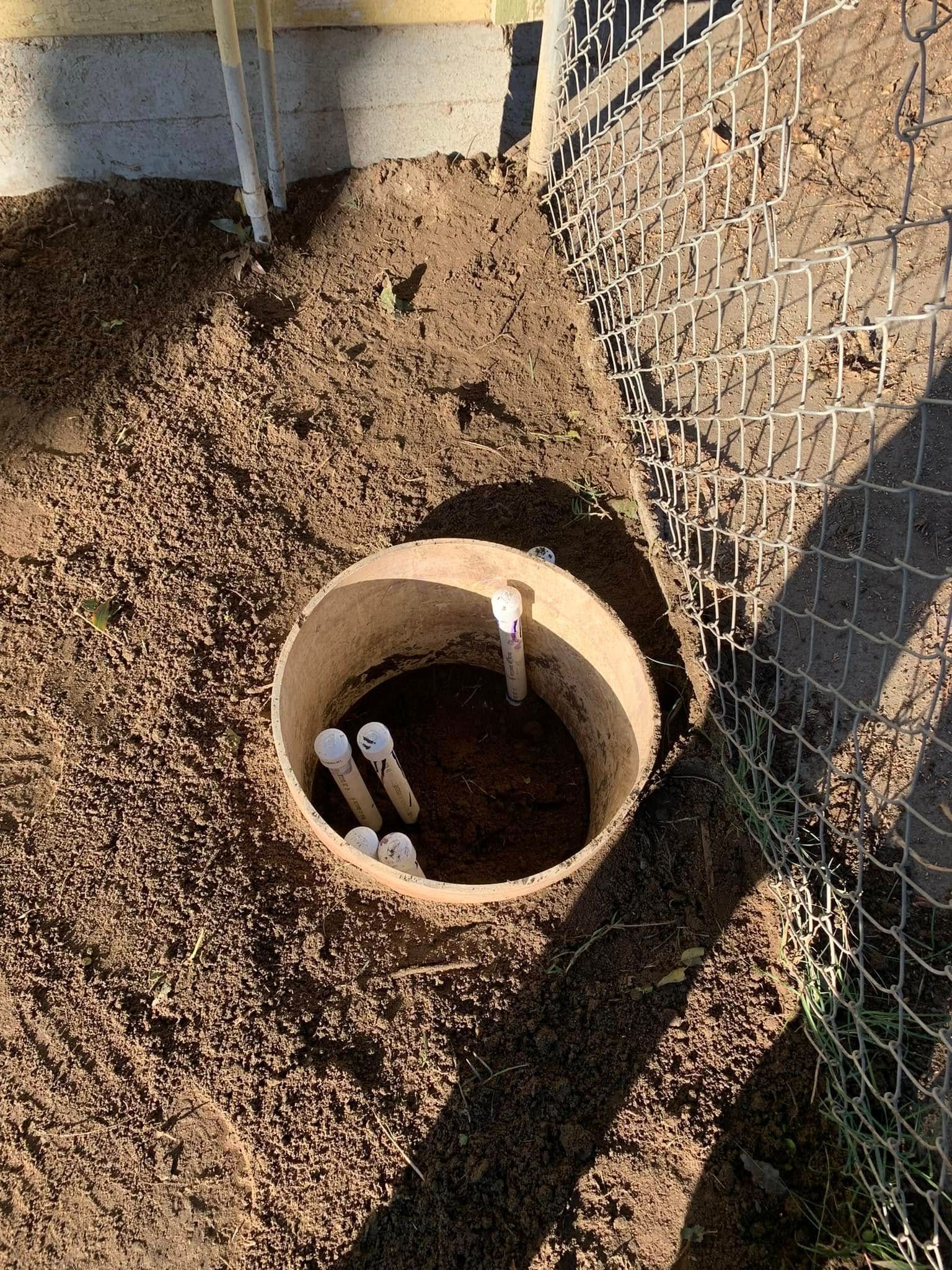 Open concrete pipe in soil beside a chain-link fence, with white pipes inside.