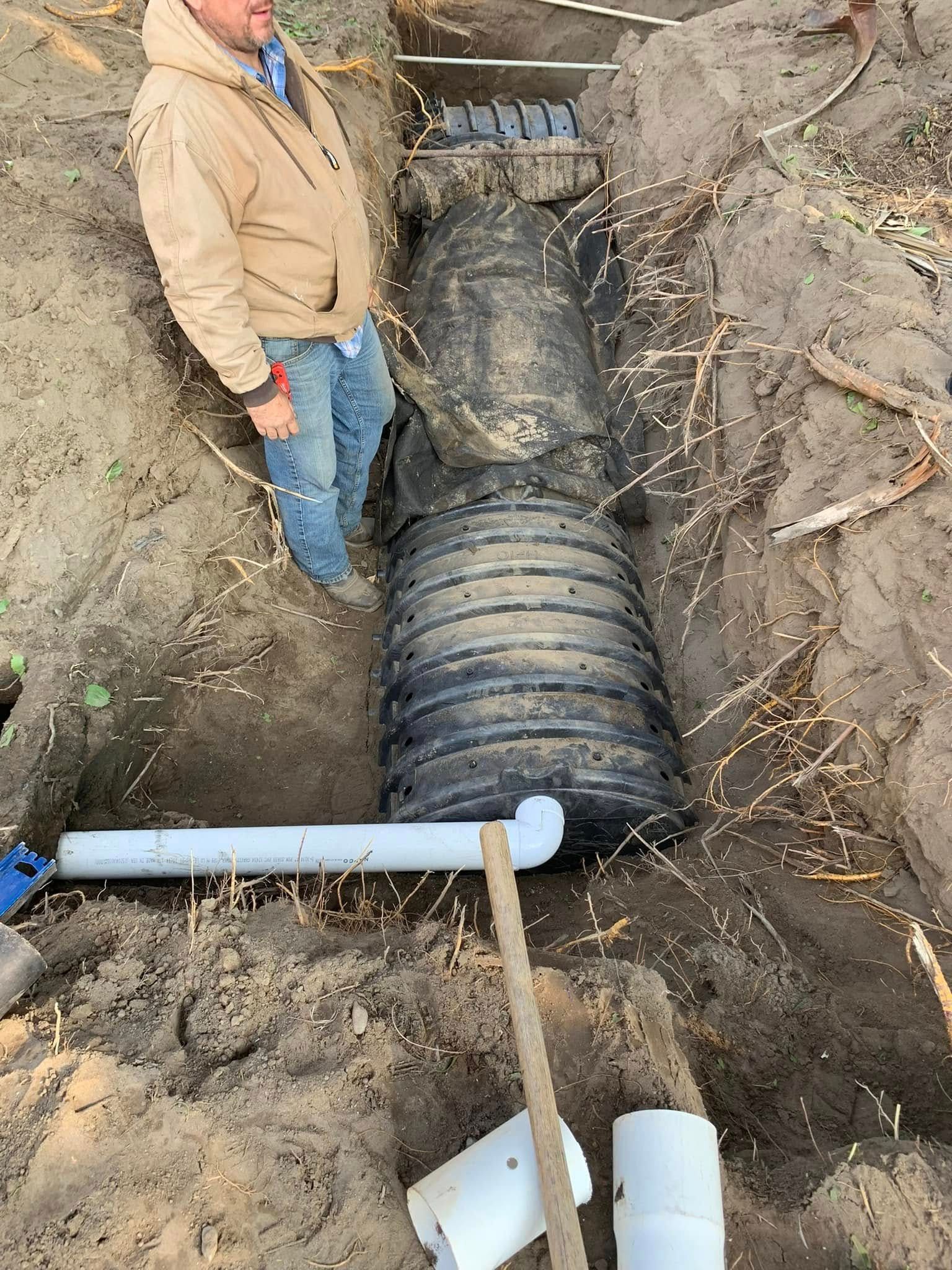 Worker beside large black pipe in a trench with dirt and exposed roots.