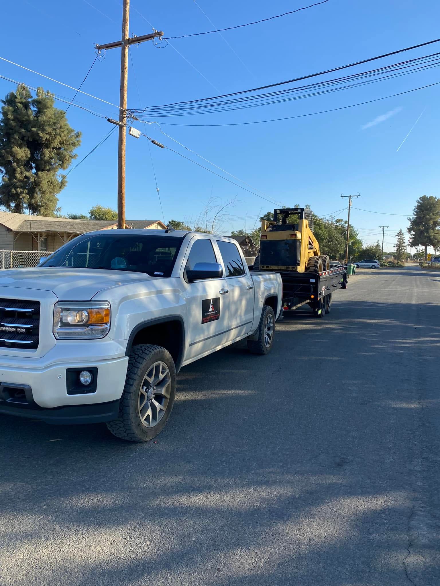 White pickup towing a yellow skid steer on a flatbed trailer along a sunny residential street