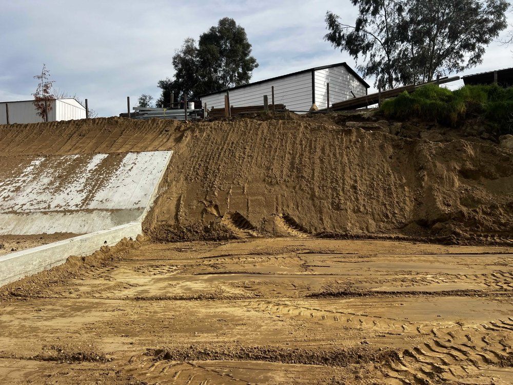 Eroded dirt embankment beside a concrete retaining wall, with a building and trees above.