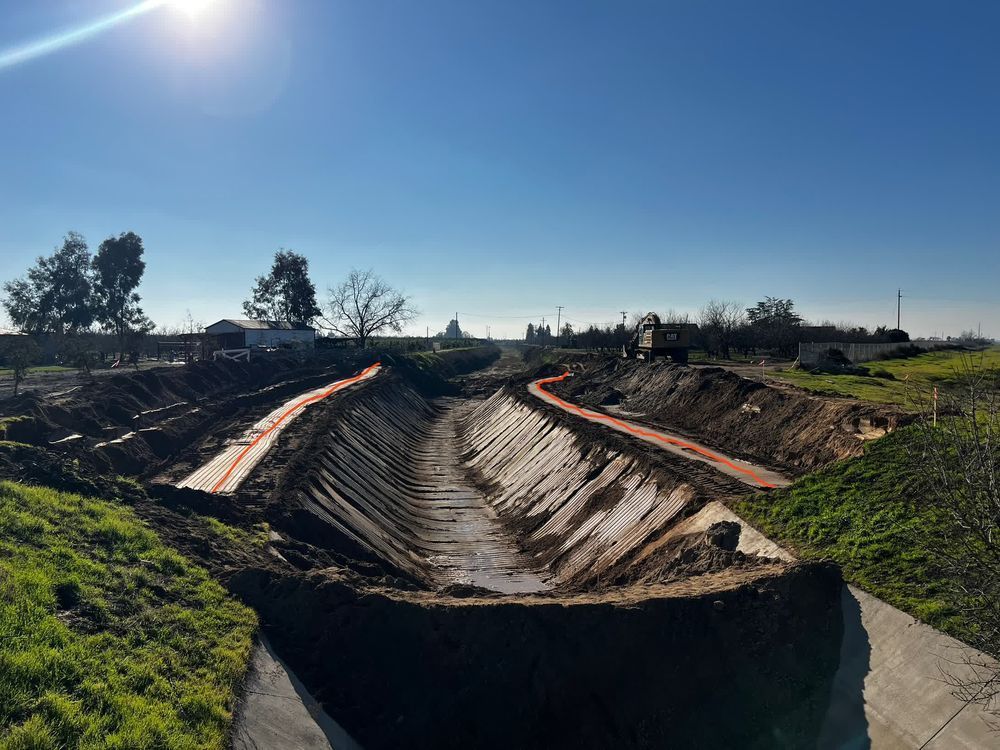 Canal under construction with concrete lining, surrounded by dirt embankments under a bright blue sky