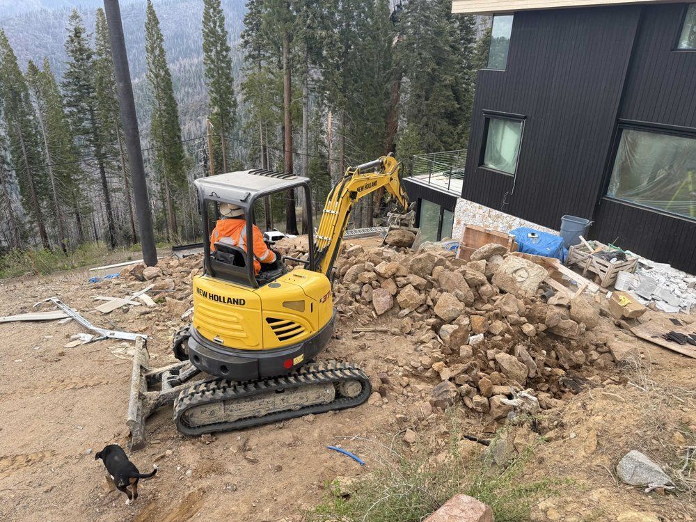 Yellow mini excavator digging beside a house, with a pile of rocks and debris nearby.