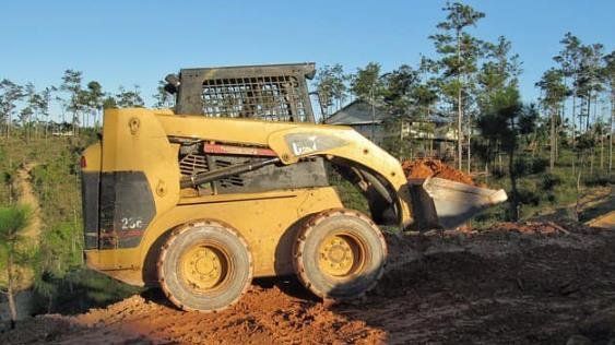 Yellow skid-steer loader moving dirt on a muddy site with trees in the background