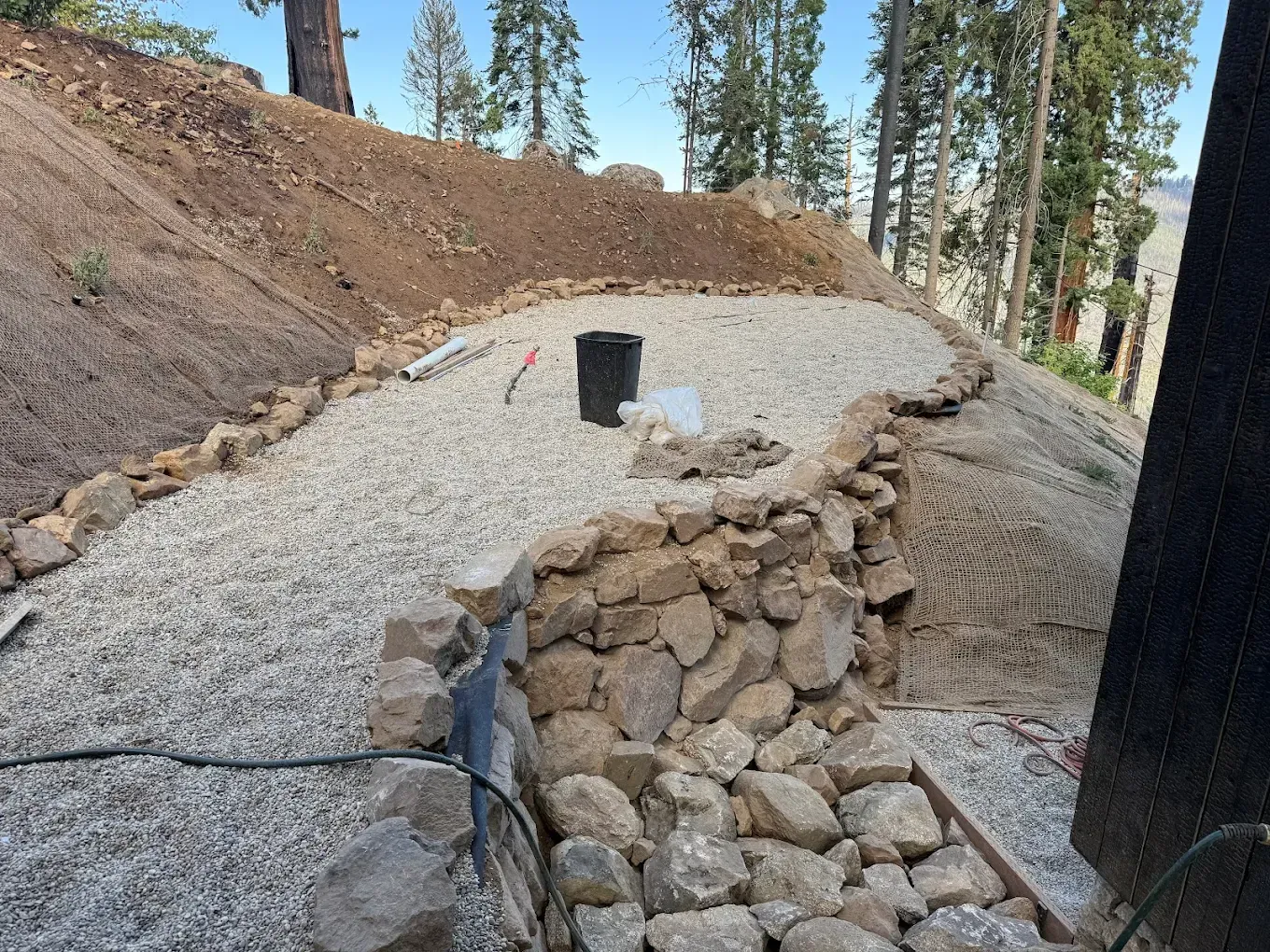 Gravel path under construction beside a dirt slope, with stone retaining wall and a black bucket.