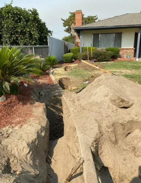 Backyard under construction with large dirt mounds, a trench, and a house in the background