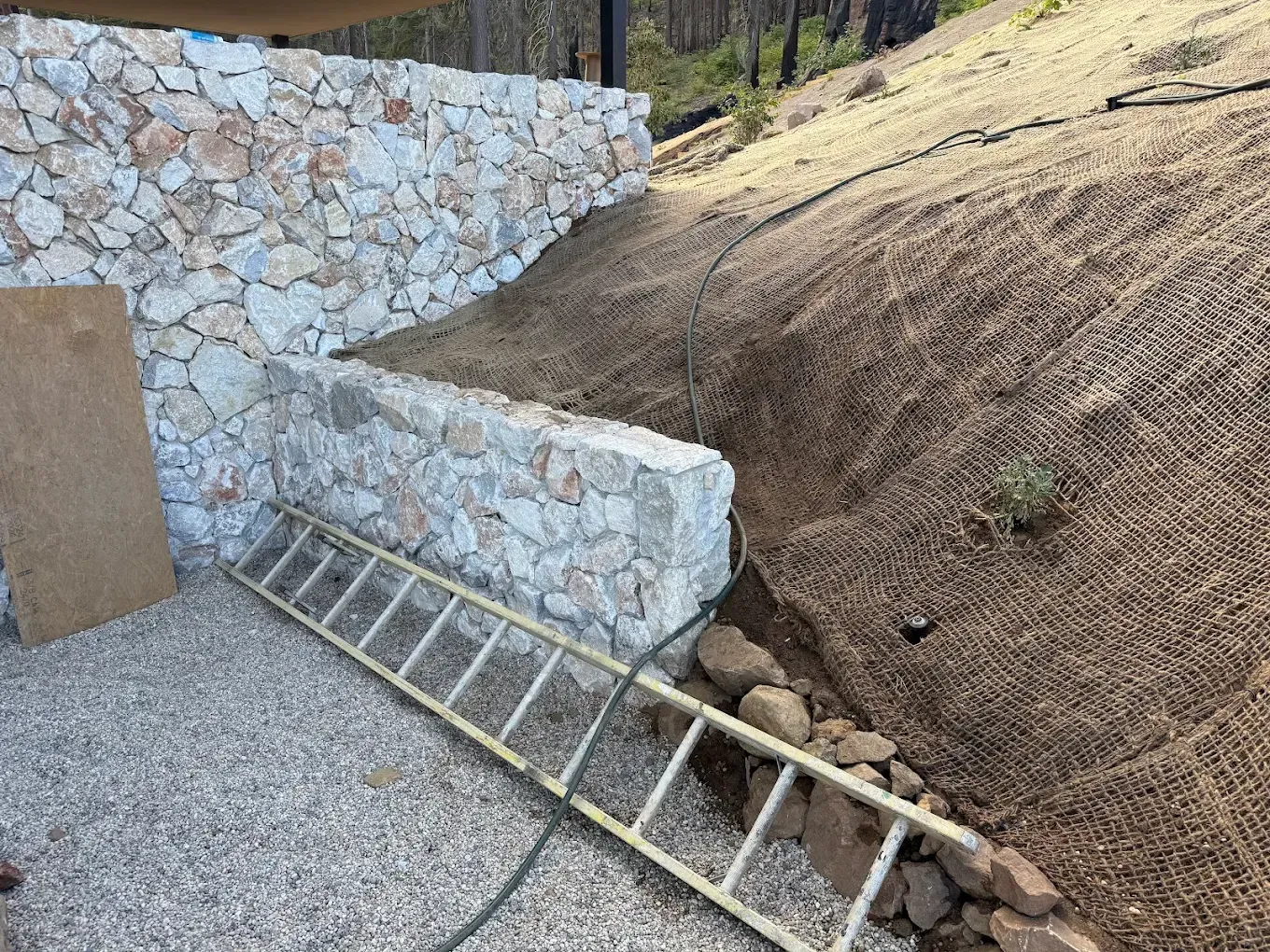 Metal ladder on gravel beside a stone retaining wall and dirt slope on a hillside.
