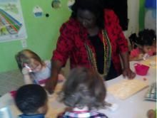 A woman in a red shirt is standing next to a group of children sitting at a table.