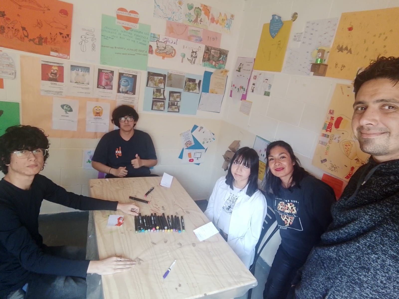 Cinco personas sentadas alrededor de una mesa, con dibujos en la pared. Caras sonrientes y pulgares arriba.