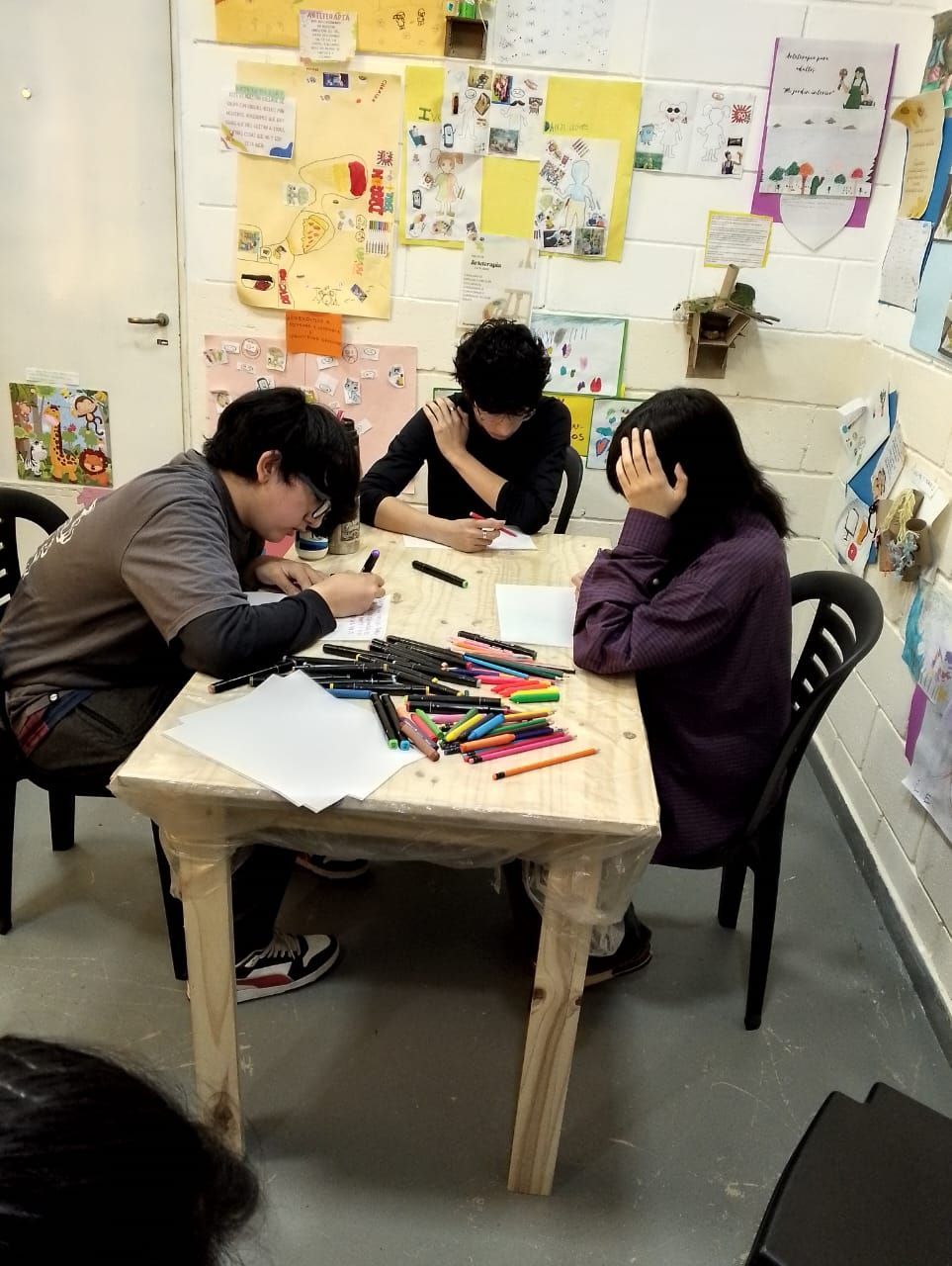 Tres personas se sientan en una mesa de madera clara, dibujando con rotuladores. Una obra de arte decora la pared detrás de ellas.