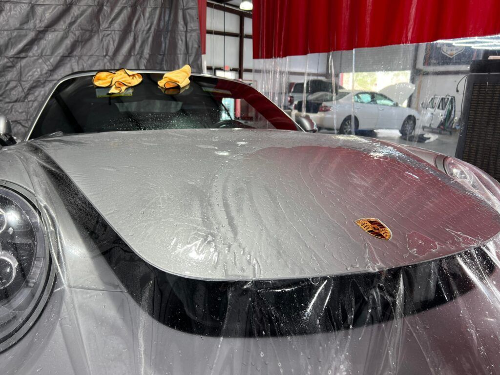 The hood of a silver porsche is covered in plastic.