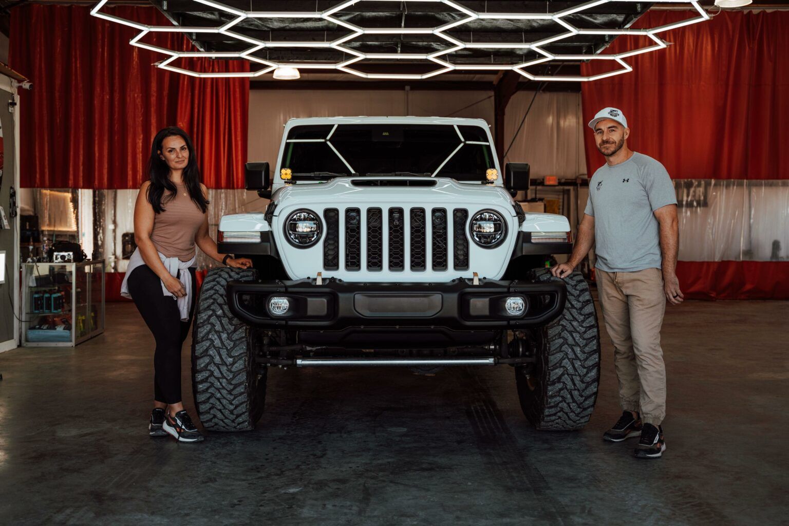 A man and a woman are standing next to a white jeep in a garage.