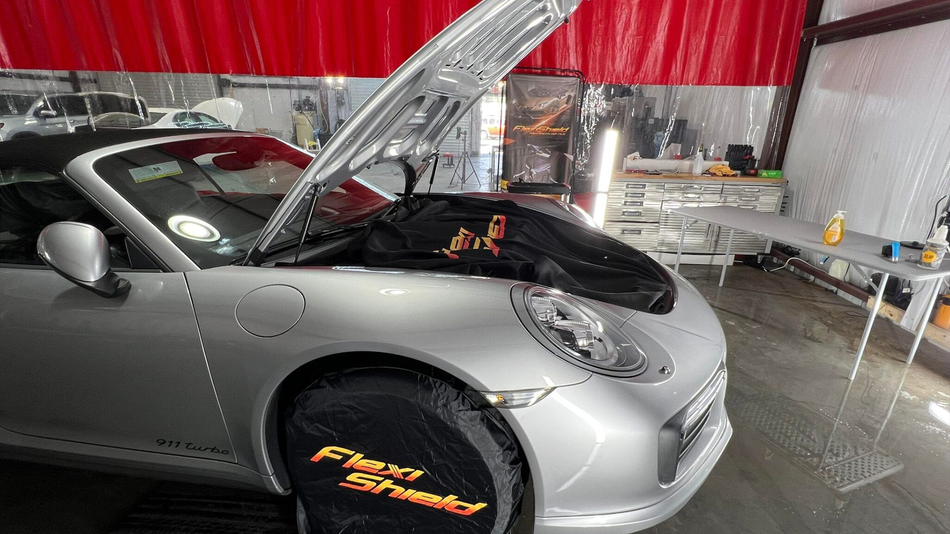 A silver porsche 911 convertible with its hood up in a garage.