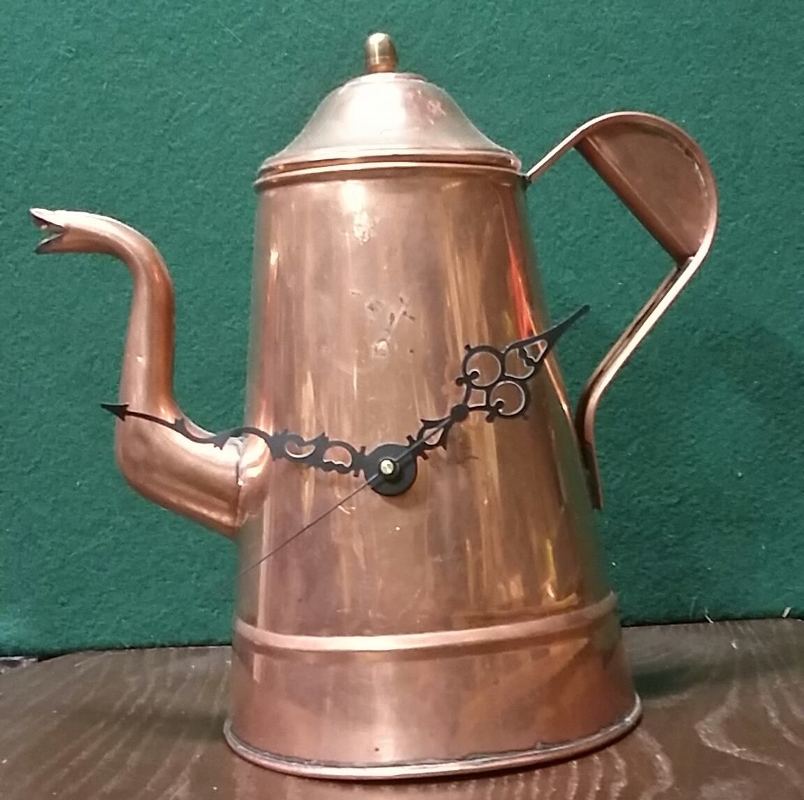 Copper teapot clock with black hands on a wooden surface against a green backdrop.